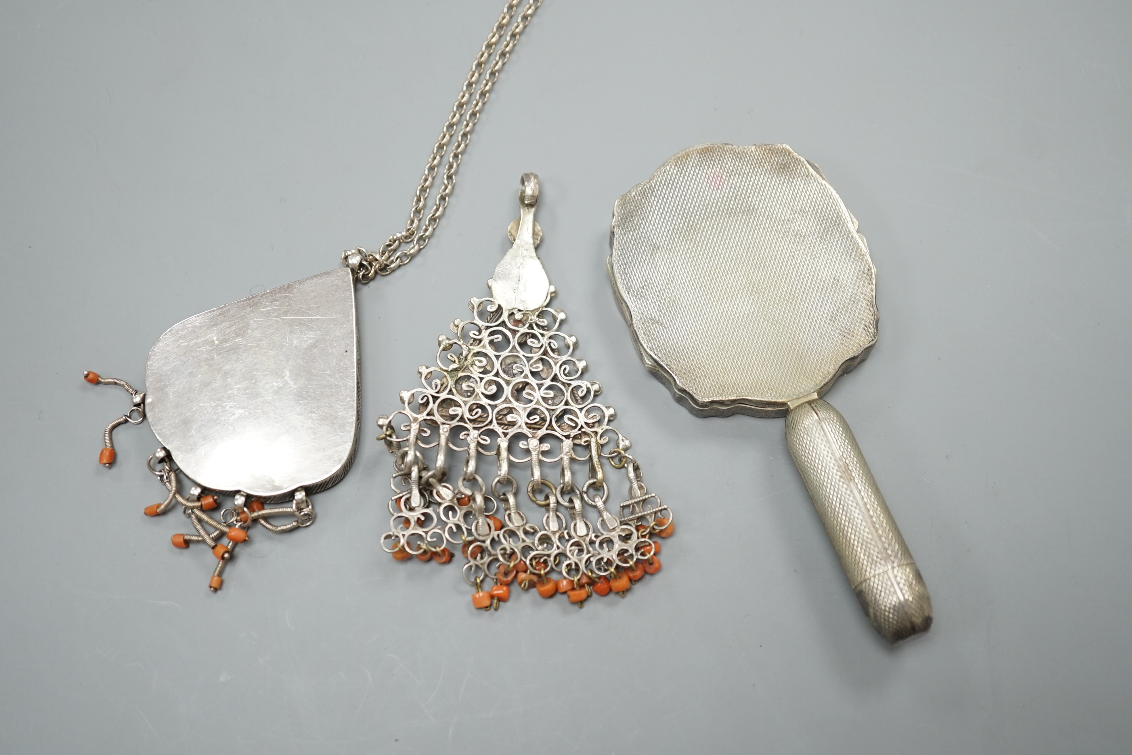 Two shaped Afghan? white metal or parcel gilt white metal, coral bead set pendants, one on a white metal chain, together with a continental engine turned white metal minaudiere, modelled as a hand mirror, 12.3cm